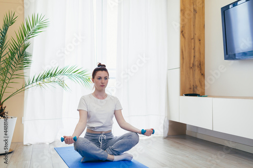 Fototapeta Naklejka Na Ścianę i Meble -  young attractive woman is practicing yoga online using a mobile phone or smartphone app. Female yogi morning in casual clothes at home in living room, workout lotus position meditates on a mat