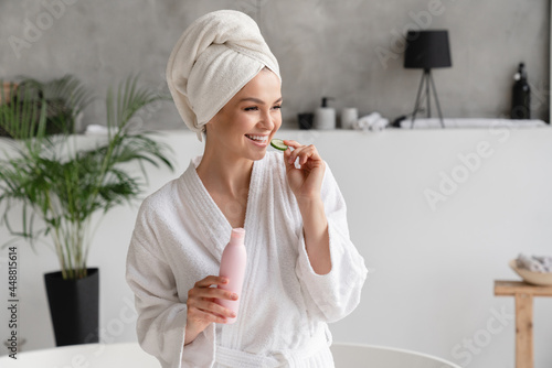 Caucasian young woman in bathrobe and spa towel eating cucumber while using beauty cosmetics for rejuvenation beautification moisturizing effect
