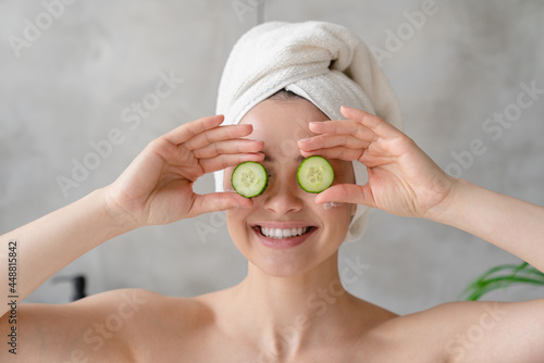 Young woman with toothy smile and spa towel holding cucumber slices on her face for rejuvenation and moisturizing effect. Beauty treatment concept