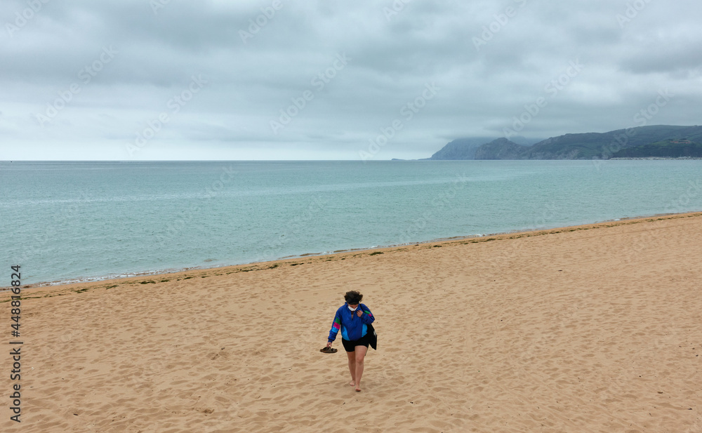 woman walks along the beach looking down on a cloudy autumn day wearing face mask woman walks along the beach looking down on a cloudy autumn day wearing face mask