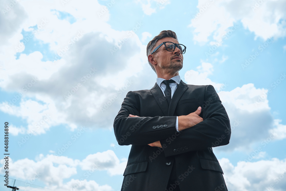 Low angle view of confident mature man in full suit keeping arms crossed Low angle view of confident mature man in full suit keeping arms crossed