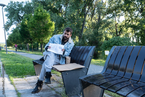 Young sick helpless desperate homeless man sitting on the bench on the urban street in the city holding help paper sign feeling hopeless and miserable hoping that someone will help him, social issues