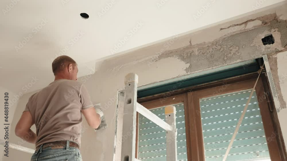Low angle view of professional construction builder aligning and smoothing plastered walls with white finishing paste solution above the window with old external roller blinds. Worker in protective