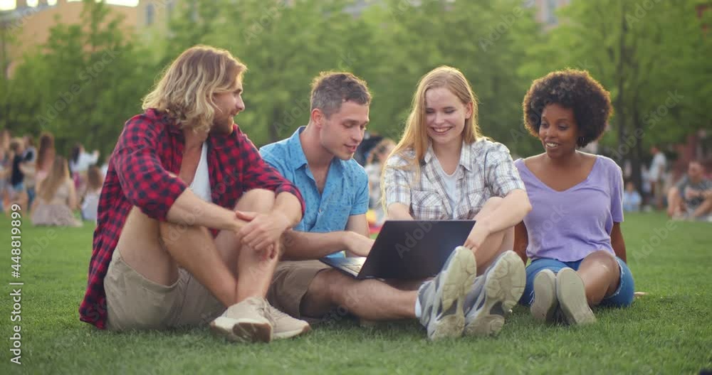 Happy young diverse friends smiling and browsing laptop while sitting on grass in park together Happy young diverse friends smiling and browsing laptop while sitting on grass in park together