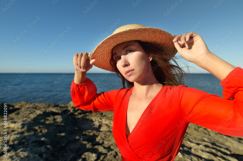 Lovely romantic caucasian young woman in red dress and straw hat against the background of the sea horizon in windy weather