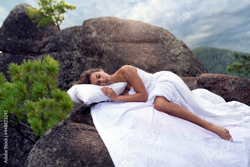 a beautiful young girl is sleeping on the rocks in the mountains  a snow-white bed  pillow and sheets are spread right on the top of the mountain  surrounded by mountains  green coniferous trees