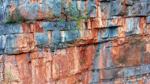 Red rugged cliff - sedimentary rock mountain formation. Vertical natural rough wall of stratum layers, colorful, sliced and cracked, brown, orange color due to ferric oxides. Geological strata canyon. photo