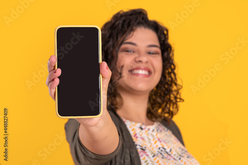 young happy brazilian woman with curly hair showing a smartphone to the camera on a yellow background