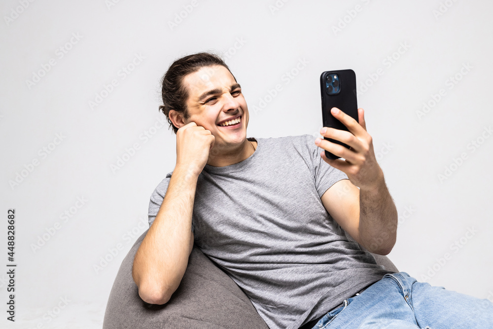 Young handsome man have video call conversation on phone sitting in bag chair isolated on white background Young handsome man have video call conversation on phone sitting in bag chair isolated on white background