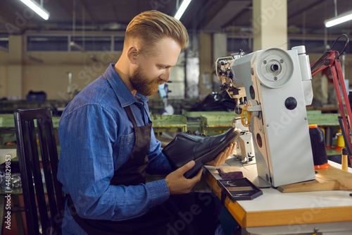 Skilled male employee of a shoe factory checks the quality of men's leather shoes at his workplace. Side view of a Caucasian bearded man sitting in a workshop with new shoes he has just made.