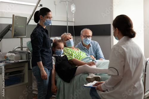 Family visiting elderly senior woman patient while wearing protective face mask in hospital ward during clinical recovery. Practitioner doctor discussing disease symptoms explaining medical treatment