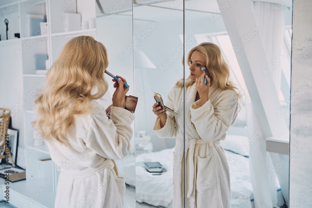 Attractive young woman in bathrobe doing make-up while looking at her reflection in the mirror - obrazy, fototapety, plakaty 