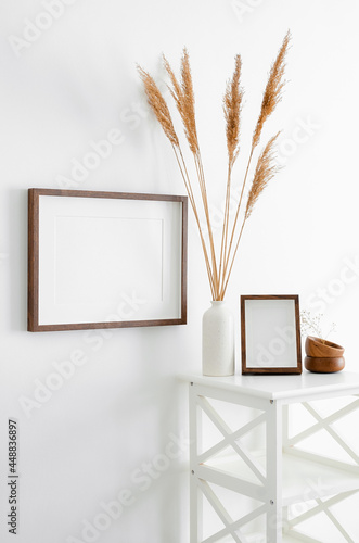 Stylish frames mockup for artwork, print or photo presentation. Blank wooden frames on white wall with furniture and ceramic vase with dry plant decorations.