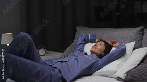 Happy woman in blue comfy pajamas going to sleep in cozy bed with soft pillows and blanket. Evening relaxation at home. End of hard day.