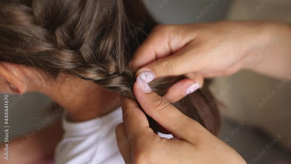 Close up shot of hands of woman weaving braid on her dark haired daughter, making hairstyle for little girl, pigtails for female kid before going to kindergarten. Close up shot of hands of woman weaving braid on her dark haired daughter, making hairstyle for little girl, pigtails for female kid before going to kindergarten.