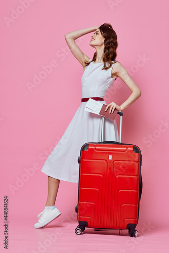 pretty woman in white dress wearing hat red suitcase passports plane tickets pink background