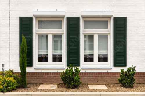 Symmetrical facade of a white modern house with an orange roof in Thorn, The Netherlands, known for its white houses and buildings. Green shutters next to the windows, greenery and plants