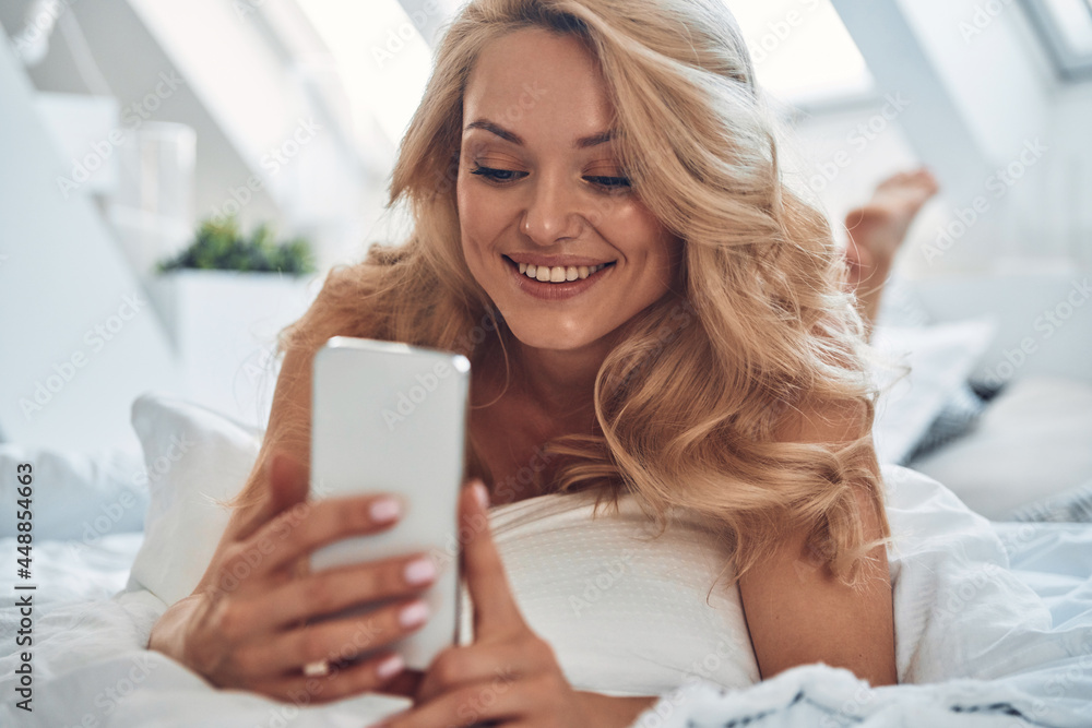 Beautiful young smiling woman using smart phone while lying in bed at home Beautiful young smiling woman using smart phone while lying in bed at home