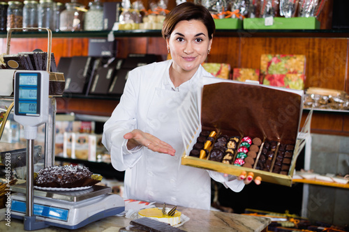 Young happy cheerful female seller offering large box of chocolates in confectionery