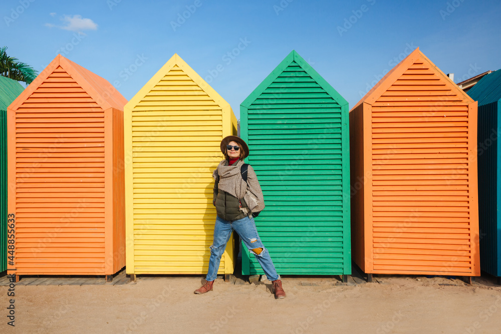 Stylish woman with a backpack and a hat on the background of beach cabins. The houses are colorful and bright.