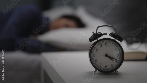 Clock standing on white table near bed with sleeping woman. Middle aged lady in pajamas having sweet dreams at night. Alarm for awaking.