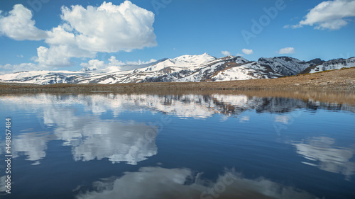 Blue sky with clouds is reflected in the mountain lake. Mountains covered with snow. Mountain landscape.