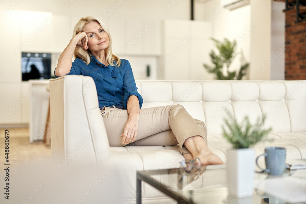Relaxed mature lady in casual wear looking at camera while sitting on a couch in modern apartment on a daytime Relaxed mature lady in casual wear looking at camera while sitting on a couch in modern apartment on a daytime