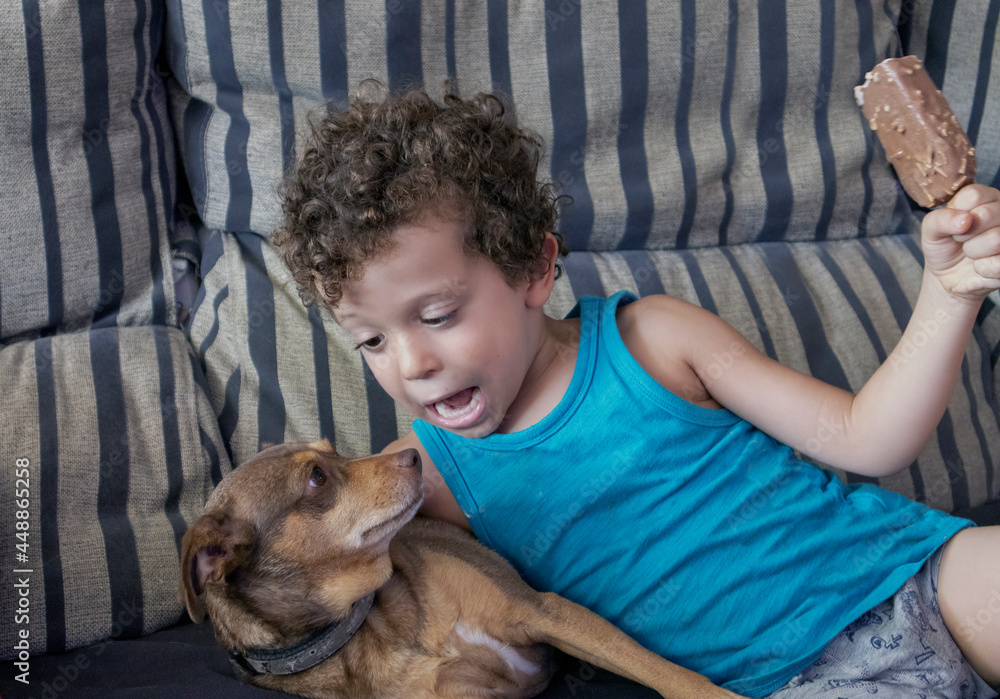 Caucasian curly-haired boy lying with his puppy on the sofa and an ice cream in his hand Caucasian curly-haired boy lying with his puppy on the sofa and an ice cream in his hand