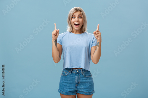 Waist-up shot of impressed enthusiastic creative blonde woman in trendy summer outfit smiling gasping being charmed and thrilled with awesome copy space pointing up over blue background