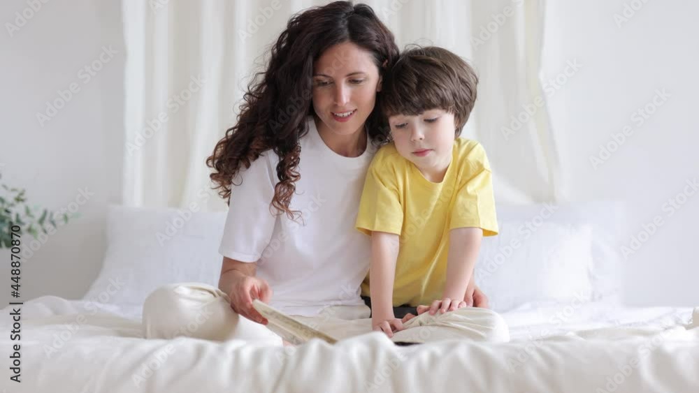 Young caring mom sit on bed with preschool son read book or show image to small kid explaining and teaching. Mother happy to spend weekend morning studying with little child at home. Family in bedroom Young caring mom sit on bed with preschool son read book or show image to small kid explaining and teaching. Mother happy to spend weekend morning studying with little child at home. Family in bedroom