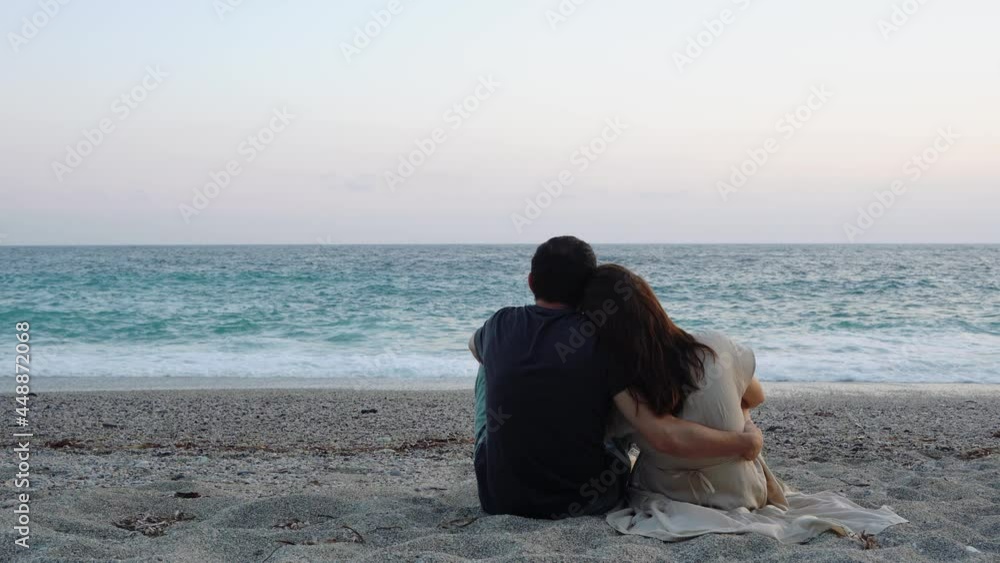 Rear view of young man hugging girlfriend while sitting on sandy beach in evening and watching beautiful seascape, lovely couple smelling fresh breeze, enjoying romantic time during summer vacation Rear view of young man hugging girlfriend while sitting on sandy beach in evening and watching beautiful seascape, lovely couple smelling fresh breeze, enjoying romantic time during summer vacation
