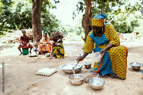Colourfully dressed black African mother preparing food for her family, with her daughters waiting in the background