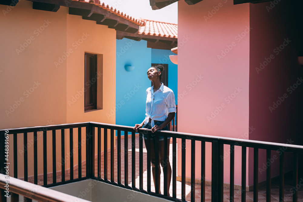 Content ethnic woman enjoying sun rays while standing at house terrace Content ethnic woman enjoying sun rays while standing at house terrace