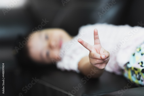 Selective focus. Happy blur child pointing up two fingers. Asian girl pointing number two fingers sign, v for victory hand gesture. Kid lying down on sofa.