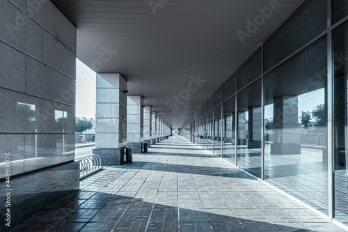 A long straight gallery in a modern building has an open side with minimalist columns and a wall made of mirror glass