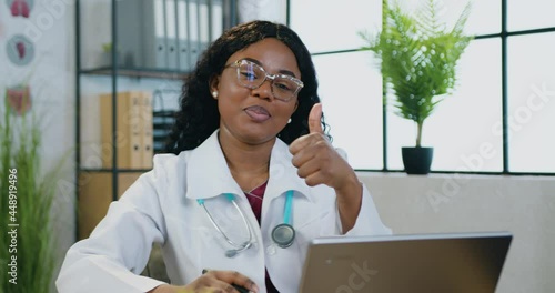 Close up of charming smiling professional cvalified black-skinned female doctor in white gawn which sitting in front of camera with computer and gesturing symbol alright photo