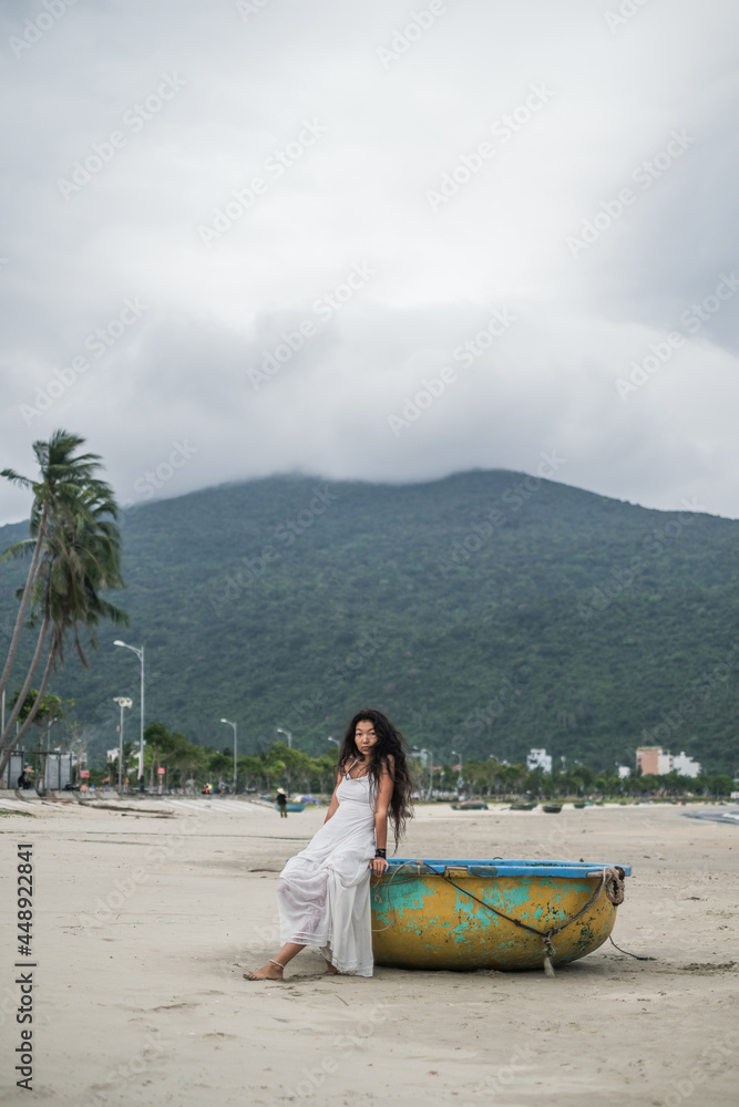 Charming young mongol woman in white dress sitting on fishing boat on the beach. Black long curly hair. Looking at camera with copy space and smiling. Romantic photo. Mountain view.