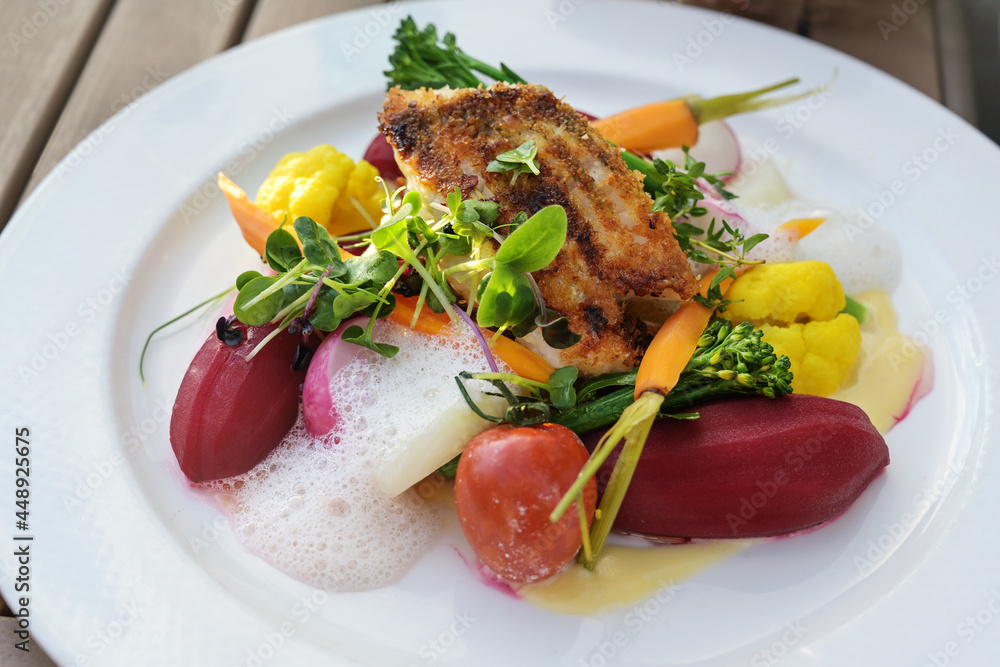 Fried fish on vegetables like carrots, cauliflower, beet and tomatoes with saffron sauce and foam, garnished with herbs and sprouts on a white plate Fried fish on vegetables like carrots, cauliflower, beet and tomatoes with saffron sauce and foam, garnished with herbs and sprouts on a white plate
