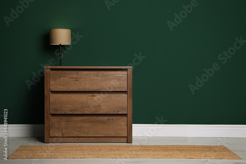 Modern wooden chest of drawers with lamp near green wall indoors. Space for text