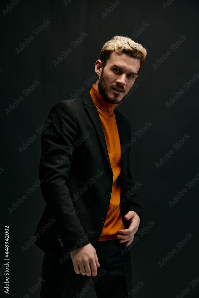 stylish man model in an orange sweater and jacket holds his hand on his belt on a dark background