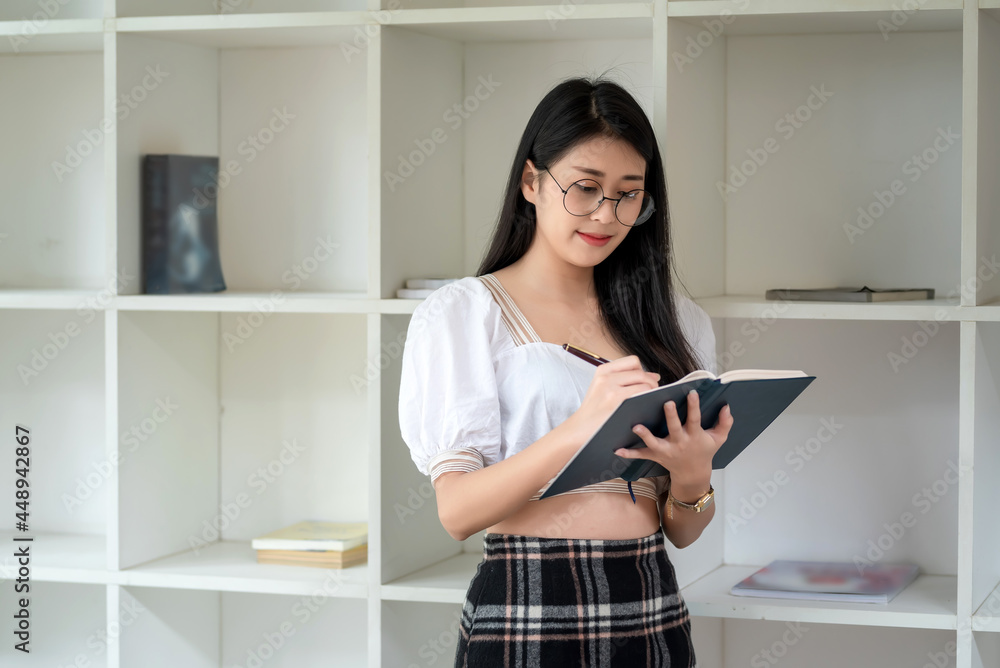 Beautiful Asian woman wearing glasses standing holding a pen and taking notes in the room. Beautiful Asian woman wearing glasses standing holding a pen and taking notes in the room.