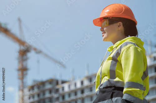 An engrossed female engineer works against the background of a multi-storey building under construction. Portrait of a young architect of a construction site, protective equipment. Selective focus.