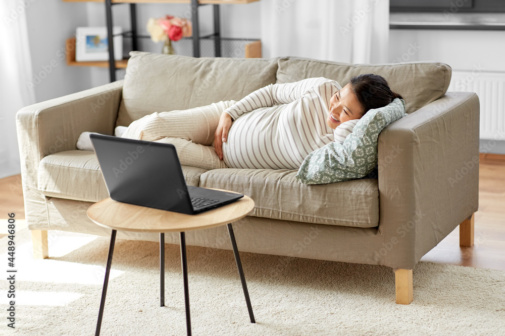 pregnancy, rest, people and expectation concept - happy smiling pregnant asian woman with laptop computer sitting on sofa at home pregnancy, rest, people and expectation concept - happy smiling pregnant asian woman with laptop computer sitting on sofa at home