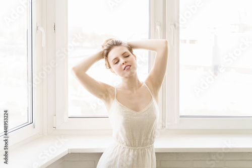 caucasian young woman in a white dress posing by the window in a bright room. morning bride