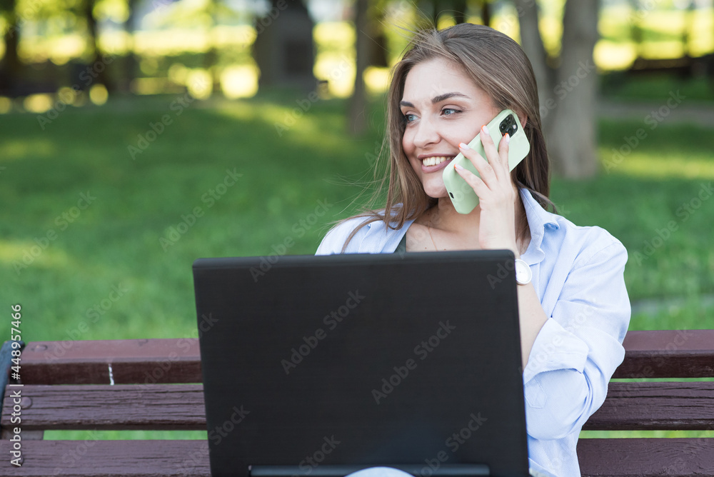 A young and attractive Caucasian girl sitting on a bench in the park. Talking on the phone and using a laptop. Freelance concept A young and attractive Caucasian girl sitting on a bench in the park. Talking on the phone and using a laptop. Freelance concept