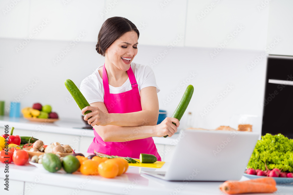 Photo of sweet funny young lady wear pink apron dancing holding cucumbers cooking dinner smiling indoors room home house