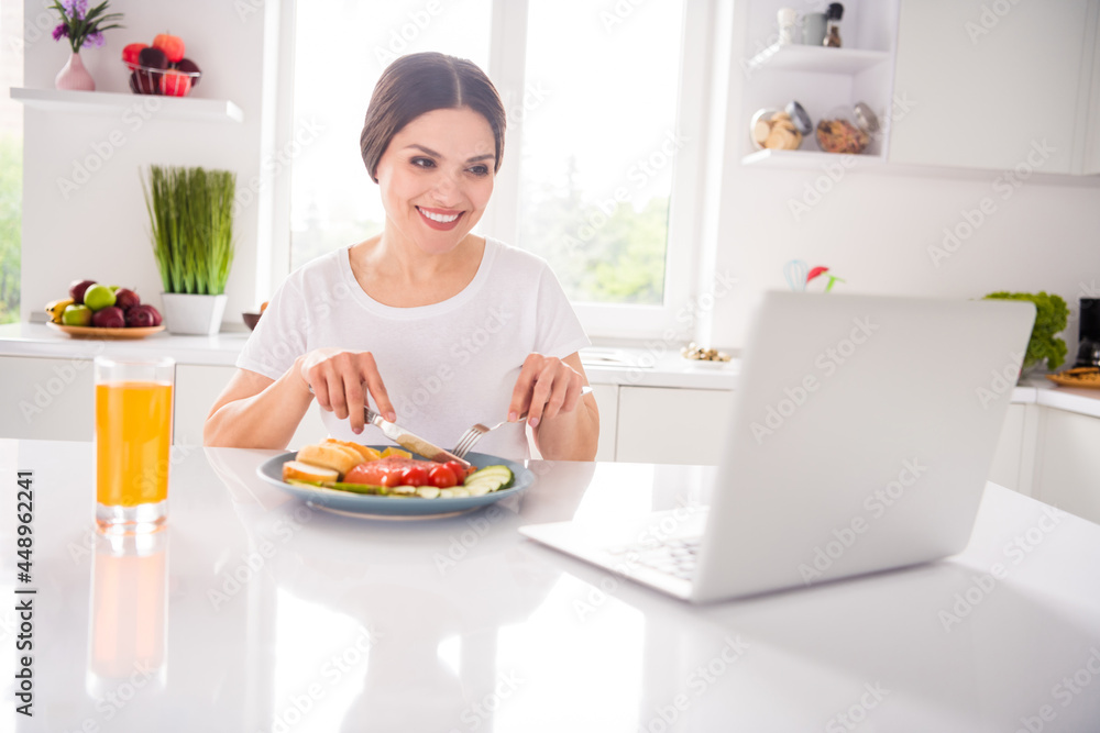 Photo of cute brunette young lady eat breakfast watch video wear t-shirt from home at table kitchen Photo of cute brunette young lady eat breakfast watch video wear t-shirt from home at table kitchen
