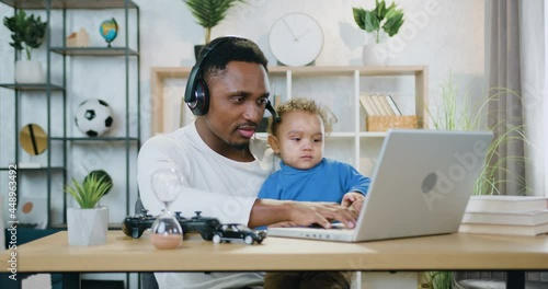 Attractive positive caring skilled black-skinned guy in headset holding his small son on knees while working on computer at home,remote work and child care concept photo