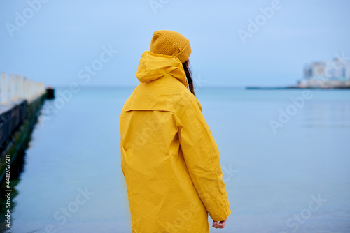 sadness woman standing alone on beach and look at distant city. loneliness woman in yellow rain coat walking coast photo