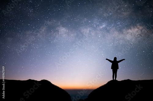 Silhouette of young female traveler and backpacker watched the star and milky way alone on top of the mountain. She raised both hands, showing enjoyed traveling and was successful.